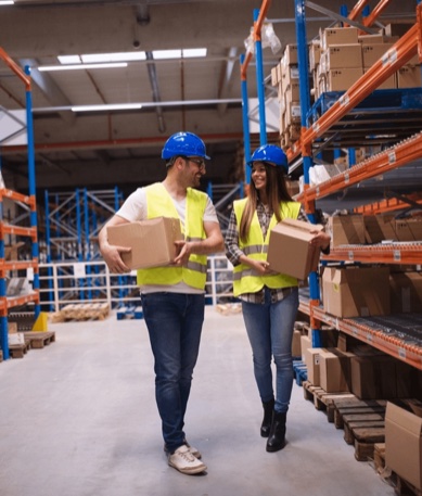 Two warehouse colleagues in hard hats and hi-vis carrying parcels beside racking