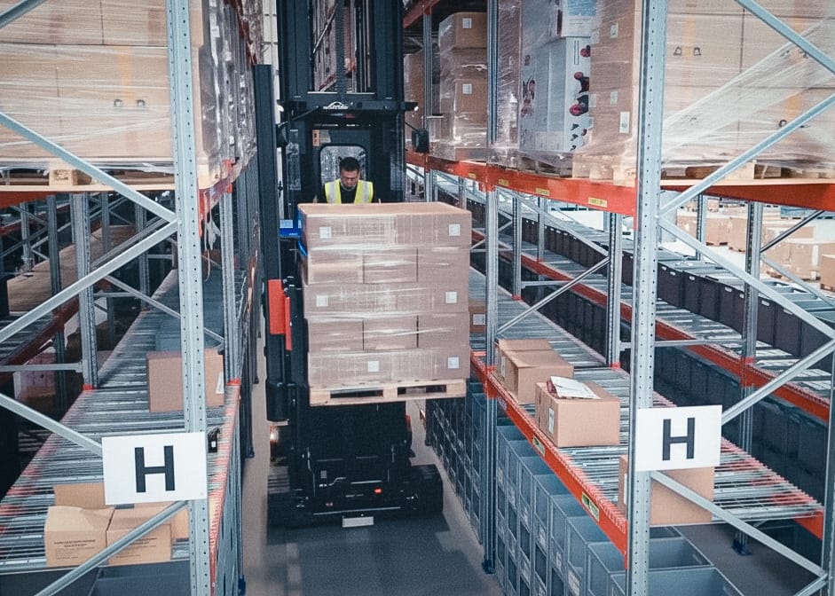 A forklift driver lifting a pallet of cartons along a tall racking aisle