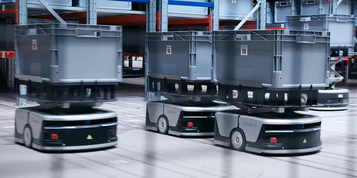 Three autonomous robots carrying grey totes across a warehouse floor